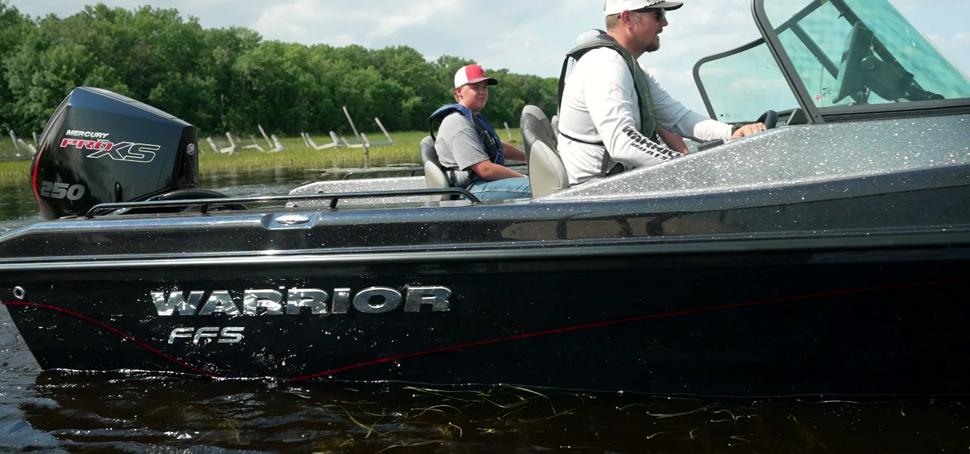 The Warrior FFS running at speed in a close broadside view, the driver at the helm and a passenger beside him, WARRIOR FFS lettering on the dark hull and a Mercury Pro XS on the transom.