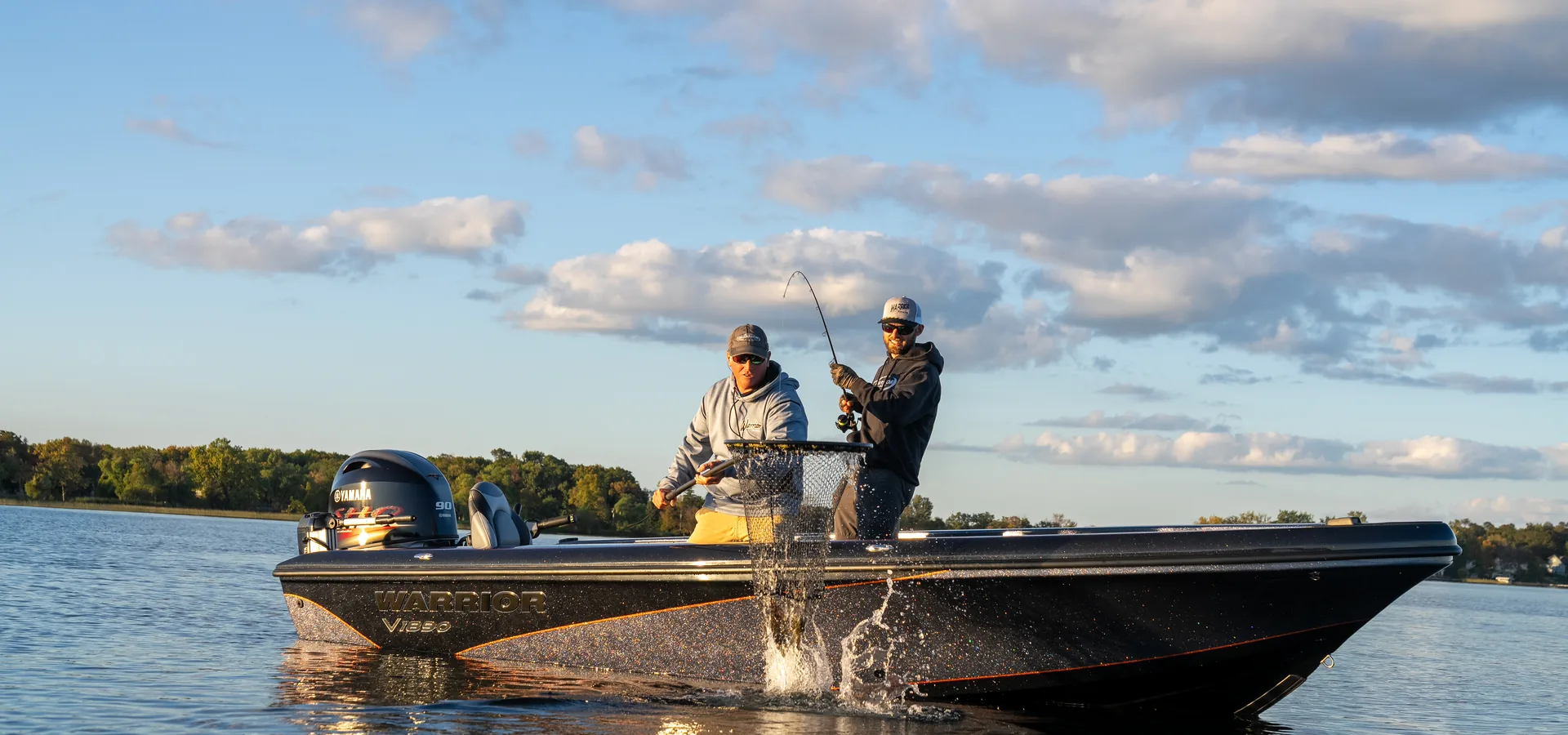 Two anglers in sweatshirts and caps net a fish from the deck of a dark blue Warrior V1890 boat on a calm lake at golden hour.