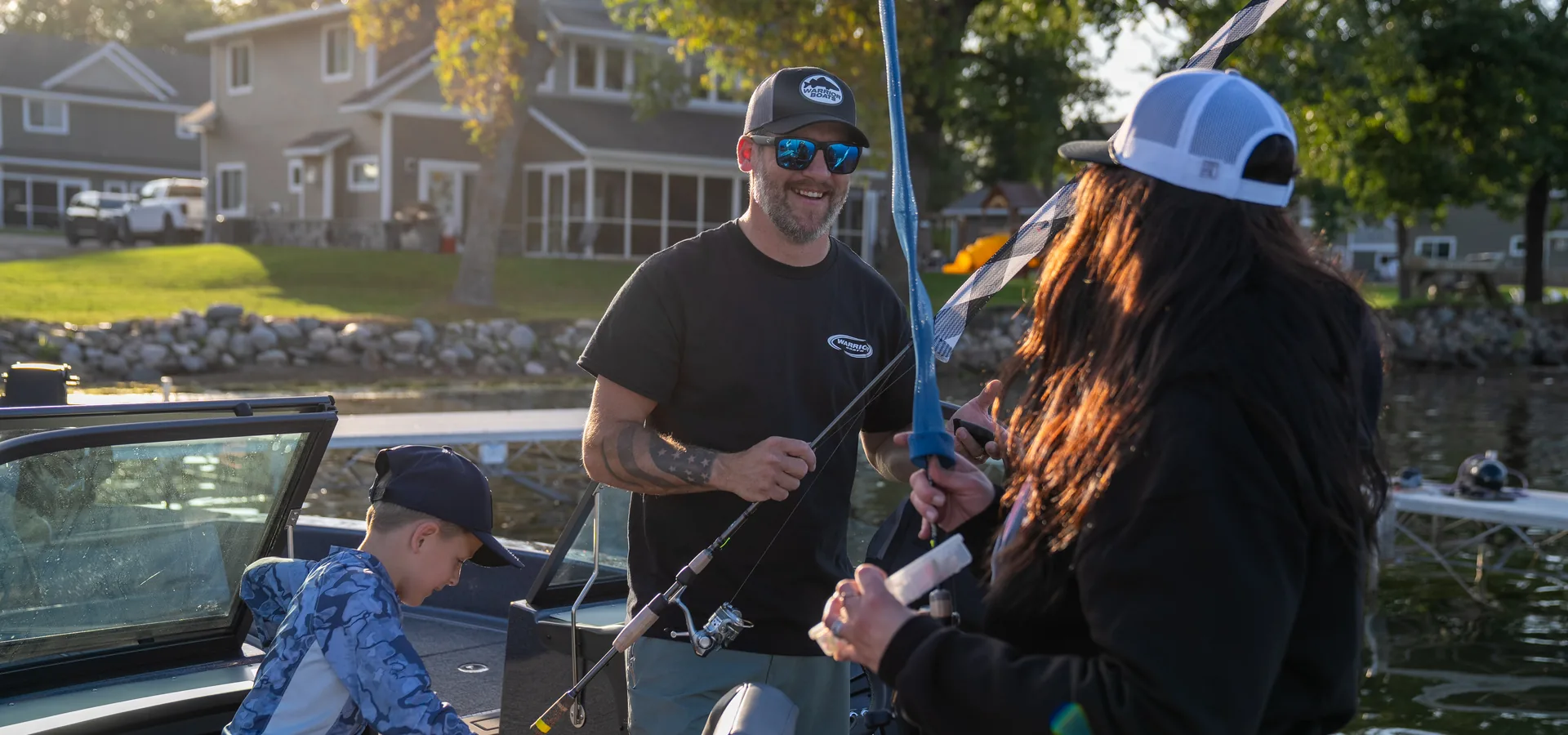 A man in a Warrior Boats cap and shirt hands a fishing rod to a woman beside a boat at a dock, with a boy in the boat and a house on the shoreline behind them.