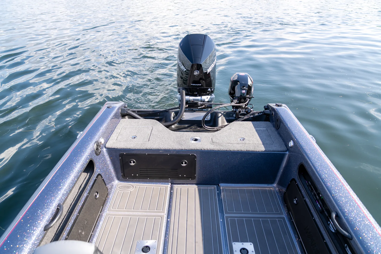 Looking down and aft over a Warrior boat's carpeted rear deck and storage hatches toward the transom, where a Mercury Verado outboard and a smaller kicker motor are mounted.