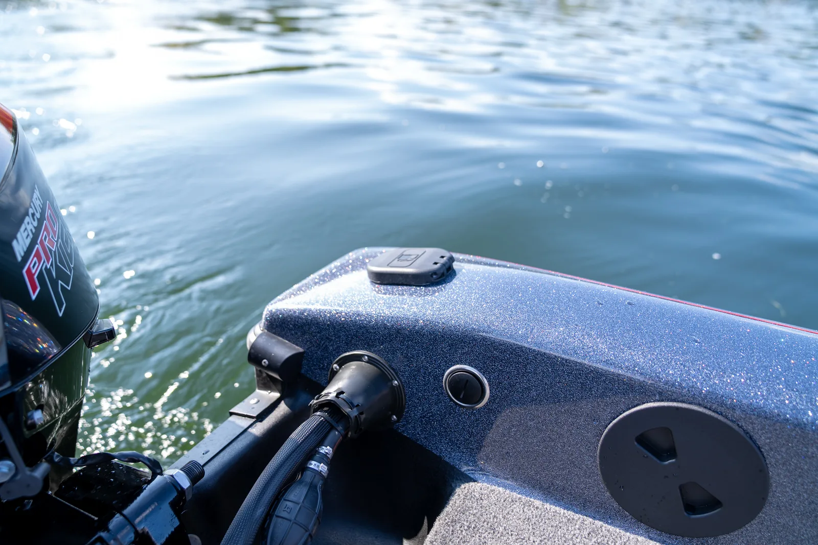 Close-up of the aft corner of a Warrior boat showing a hinged cover in the metal-flake transom cap, a hose pass-through fitting on the transom, and a round twist-off deck cap, with a Mercury Pro Kicker blurred at the left edge.