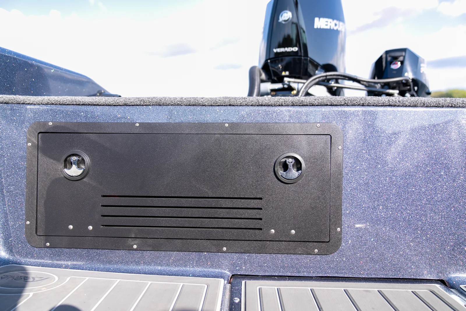 A black access panel with two flush latches and a louvered vent set into the metal-flake bulkhead below a Warrior boat's rear deck, with the Mercury outboard visible above.