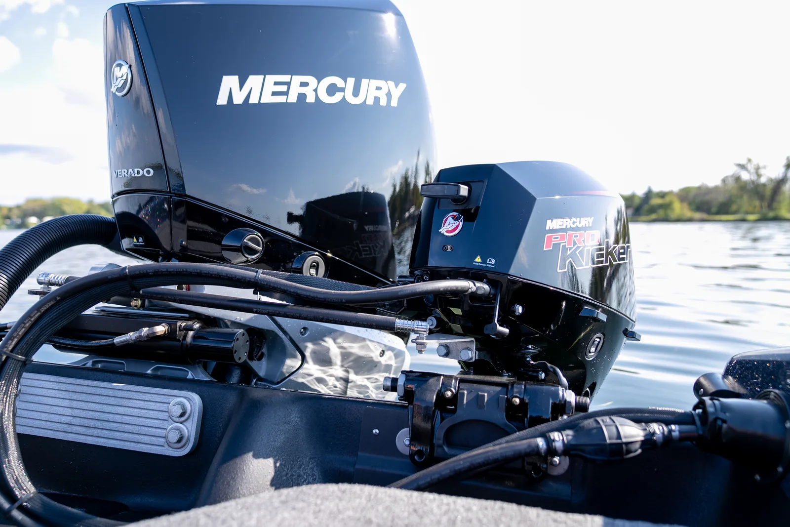 Close view of a black Mercury Verado outboard alongside a smaller Mercury Pro Kicker motor on the transom, with a tree-lined shore behind.