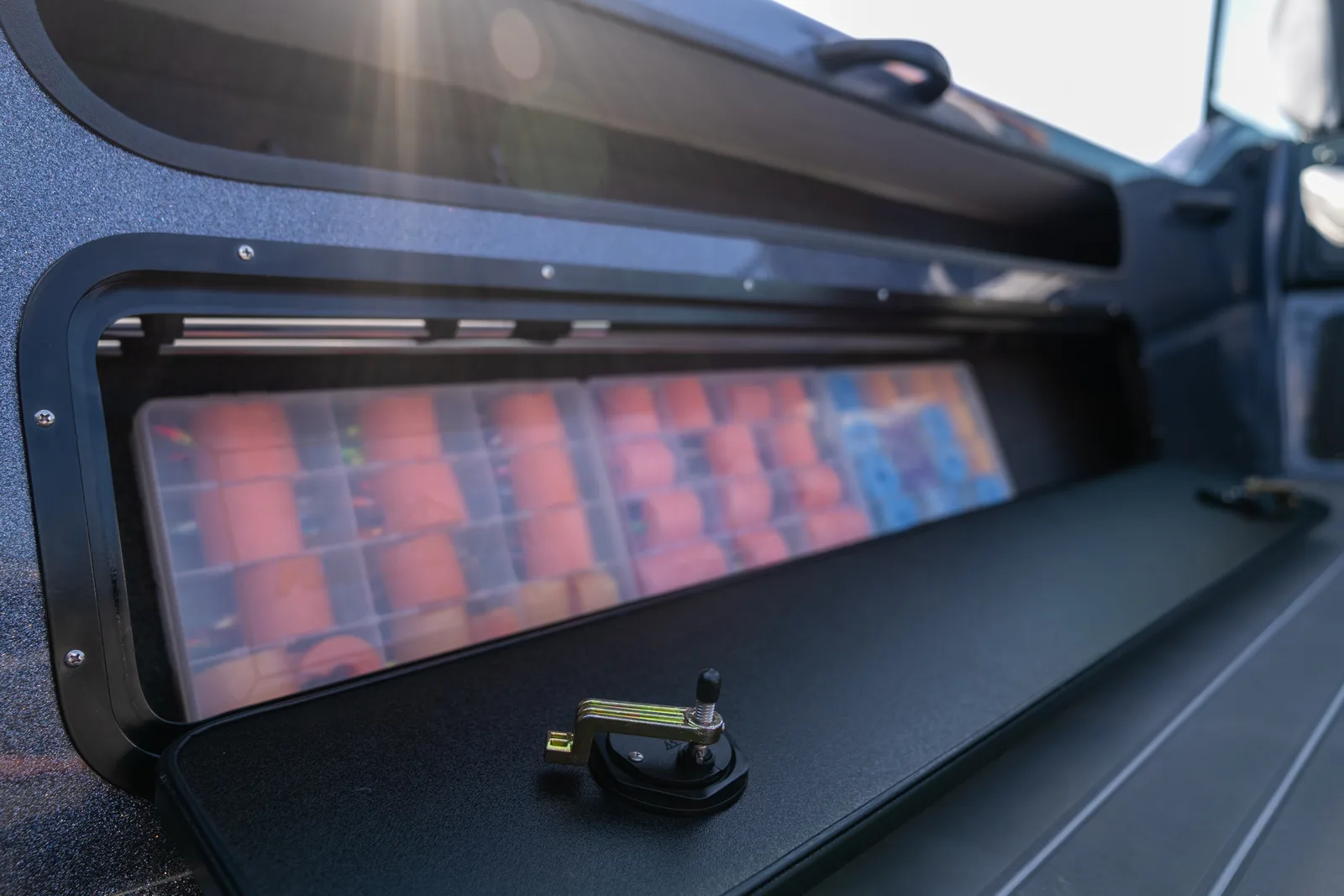 An open gunwale storage compartment on a blue metal-flake boat, its drop-down door secured by a cam lock lever, with translucent tackle trays stacked inside.