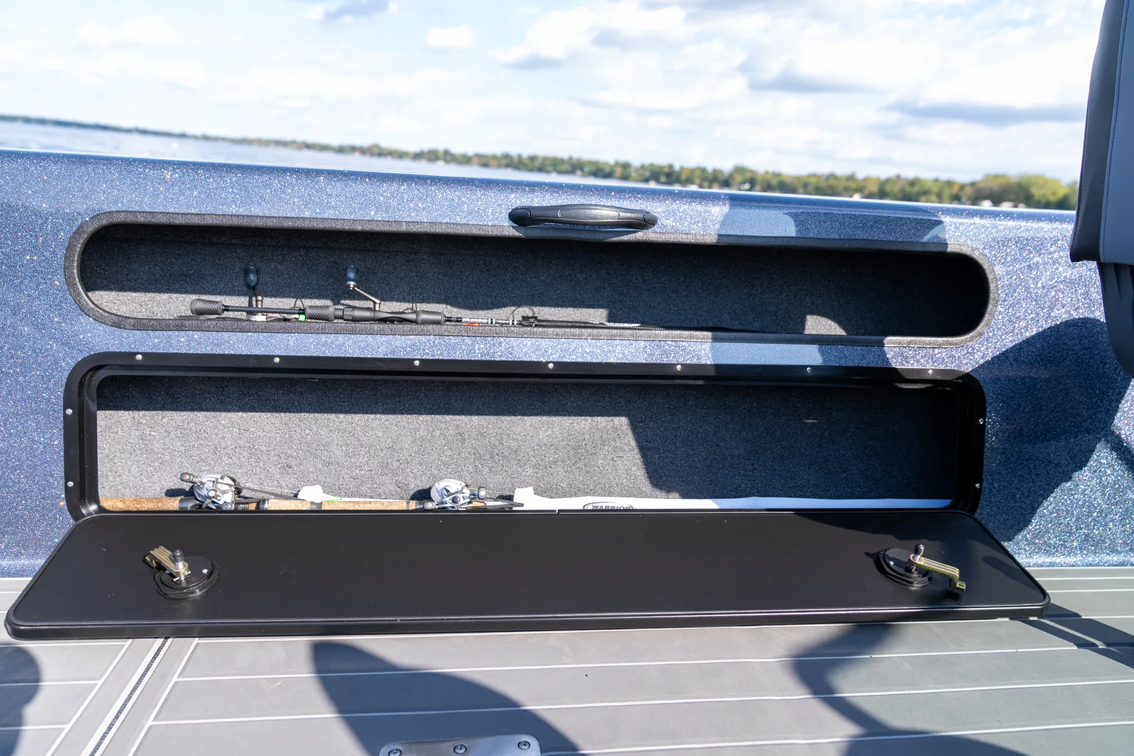 A gunwale rod locker standing open on a blue metal-flake boat, with fishing rods and baitcasting reels lying inside on grey carpet and a lake and treed shoreline behind.