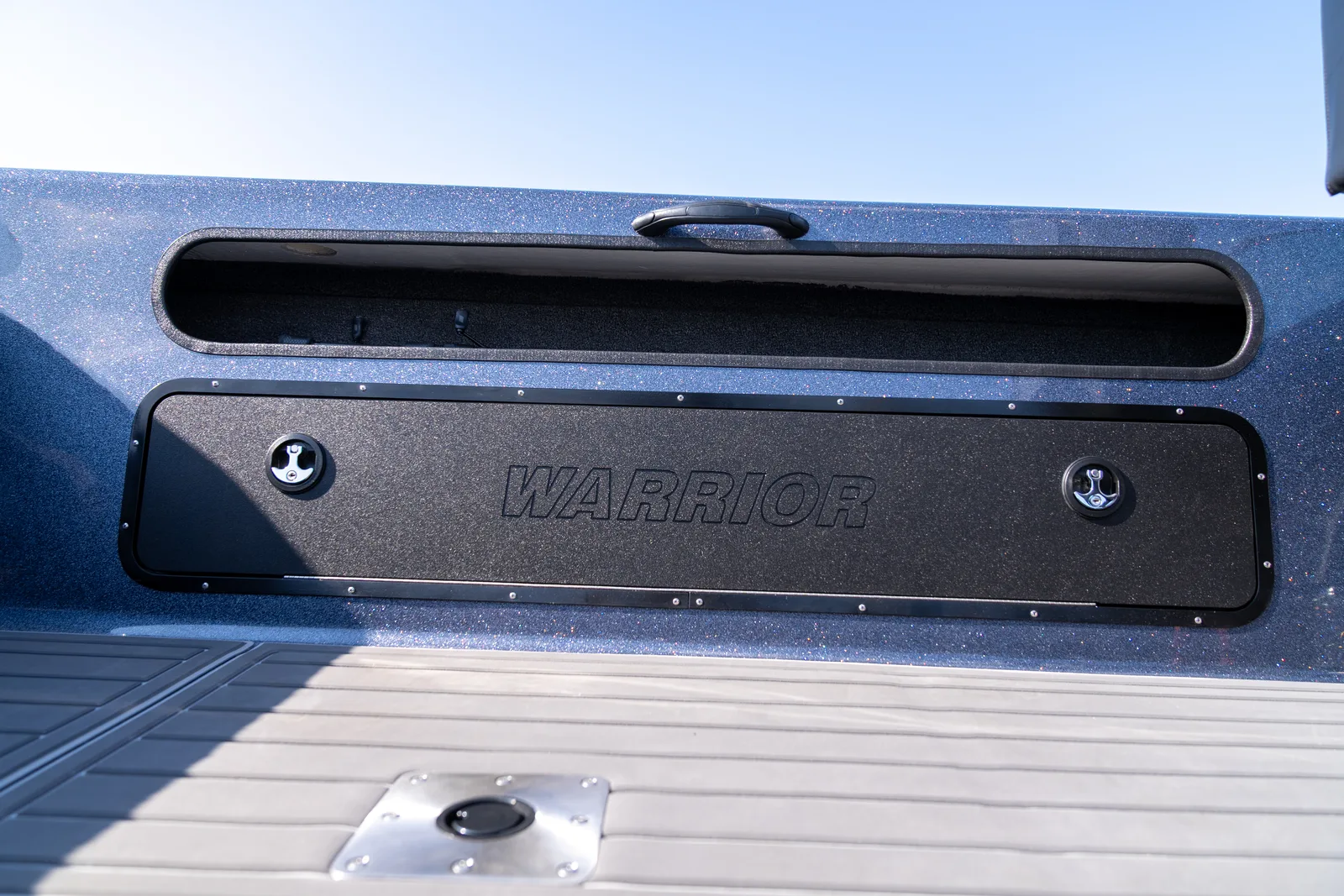 A closed black gunwale storage door embossed with the Warrior logo and secured by two flush latches, with an open carpeted rod trough above it and a stainless flush-mount seat base on the deck below.