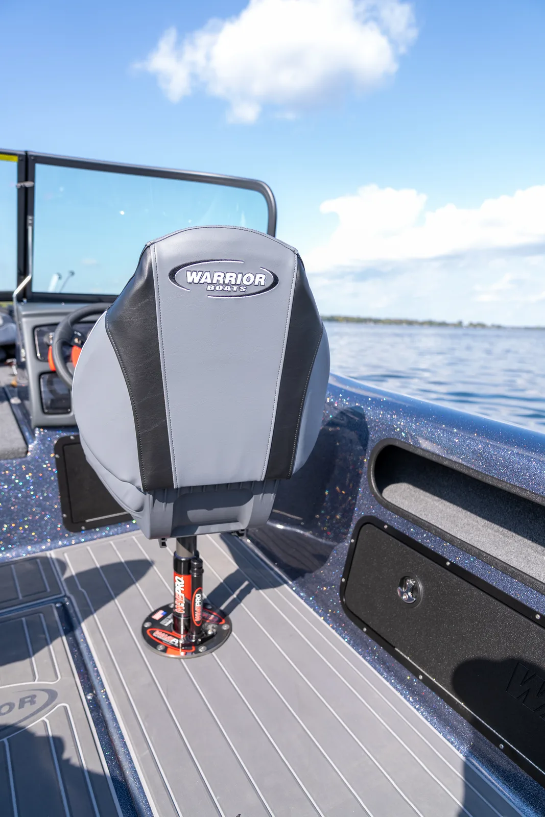 A grey and black swivel seat with a Warrior Boats logo on the backrest, mounted on a shock-absorbing pedestal on the deck, with the steering wheel and windshield beyond it and open water to the right.