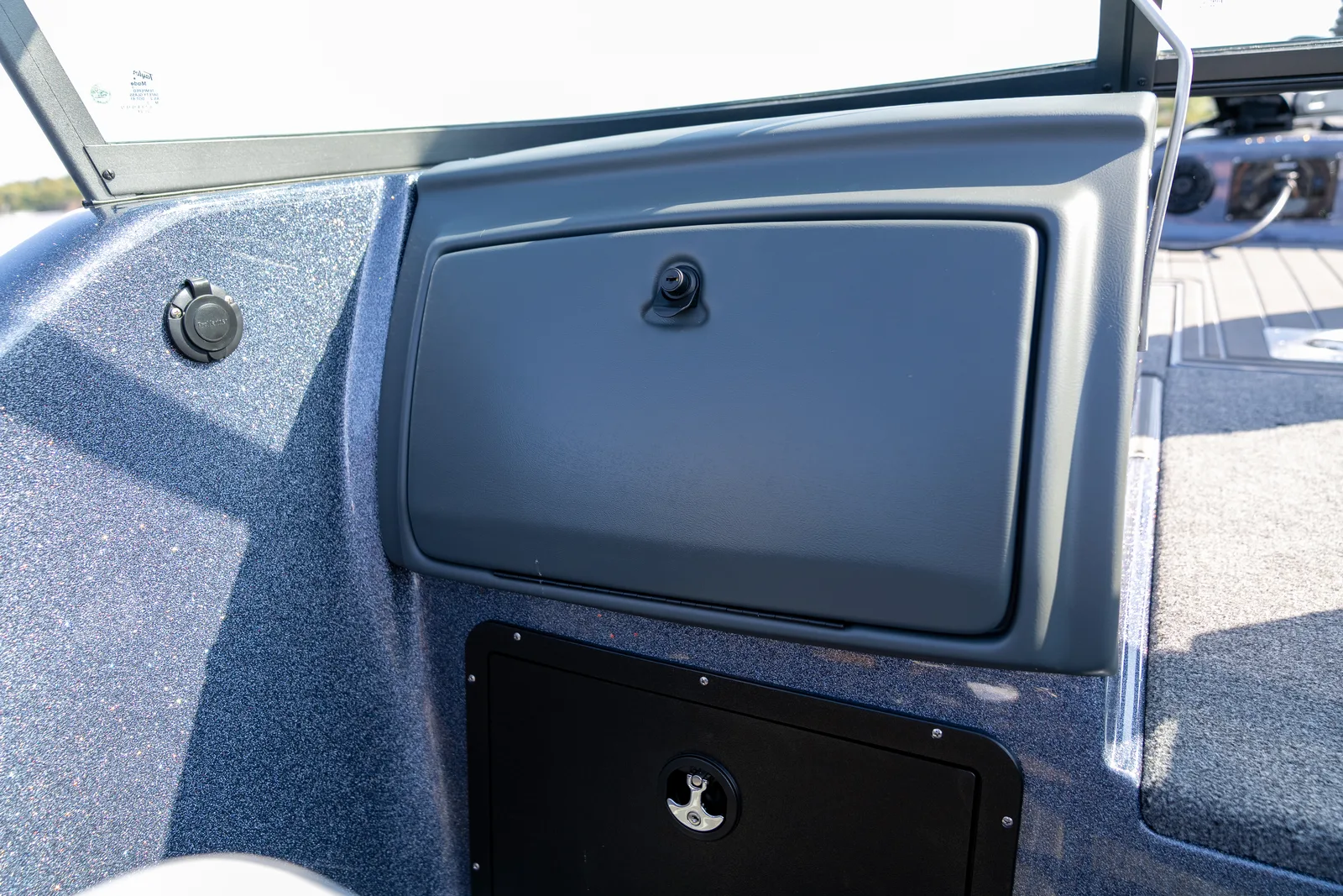 A latched grey glove box door in the console of a boat, with a windshield above and a black access hatch in the panel below.