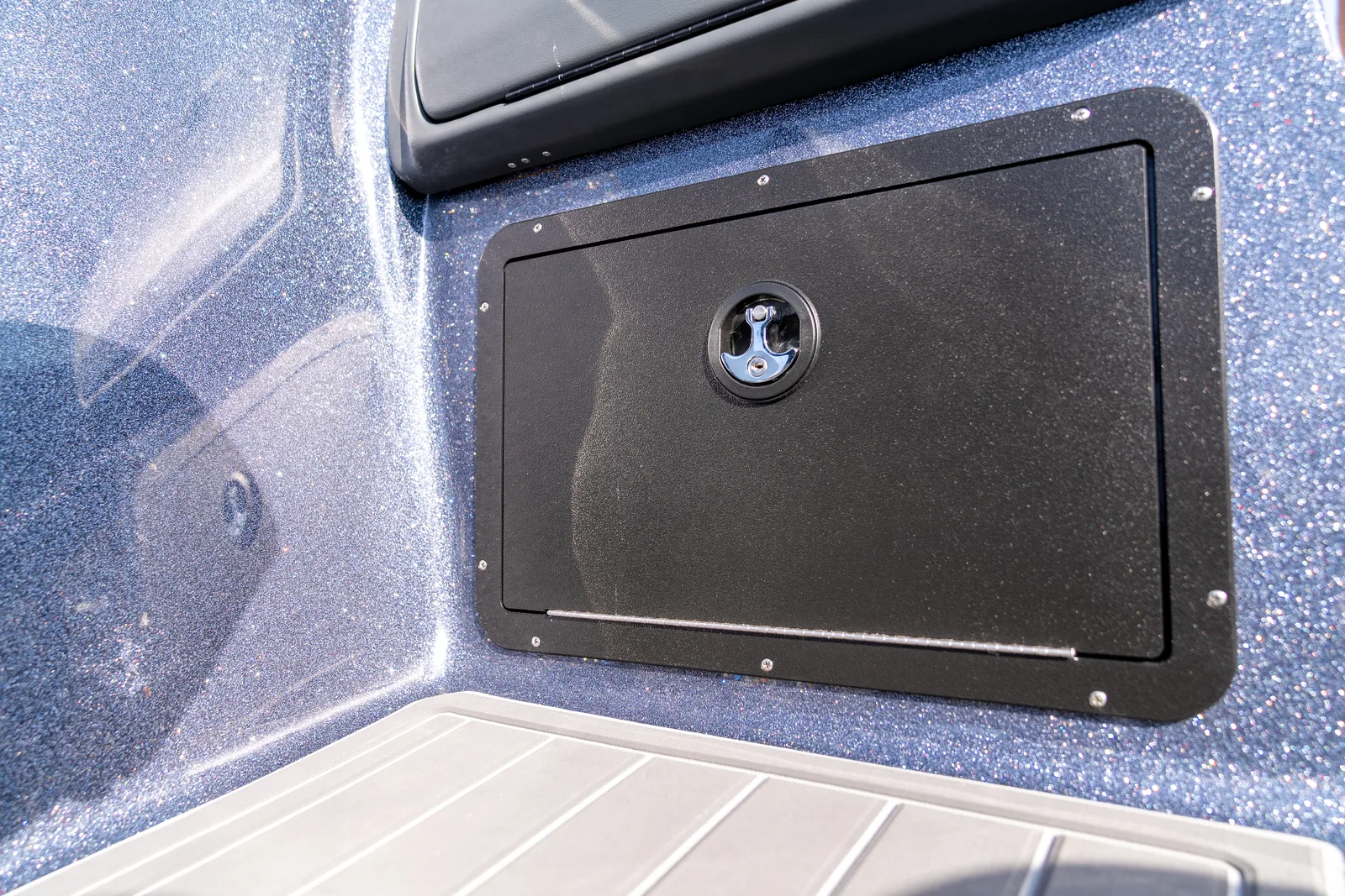 A closed black storage hatch with a chrome T-handle latch mounted in the blue metalflake side panel of a boat.