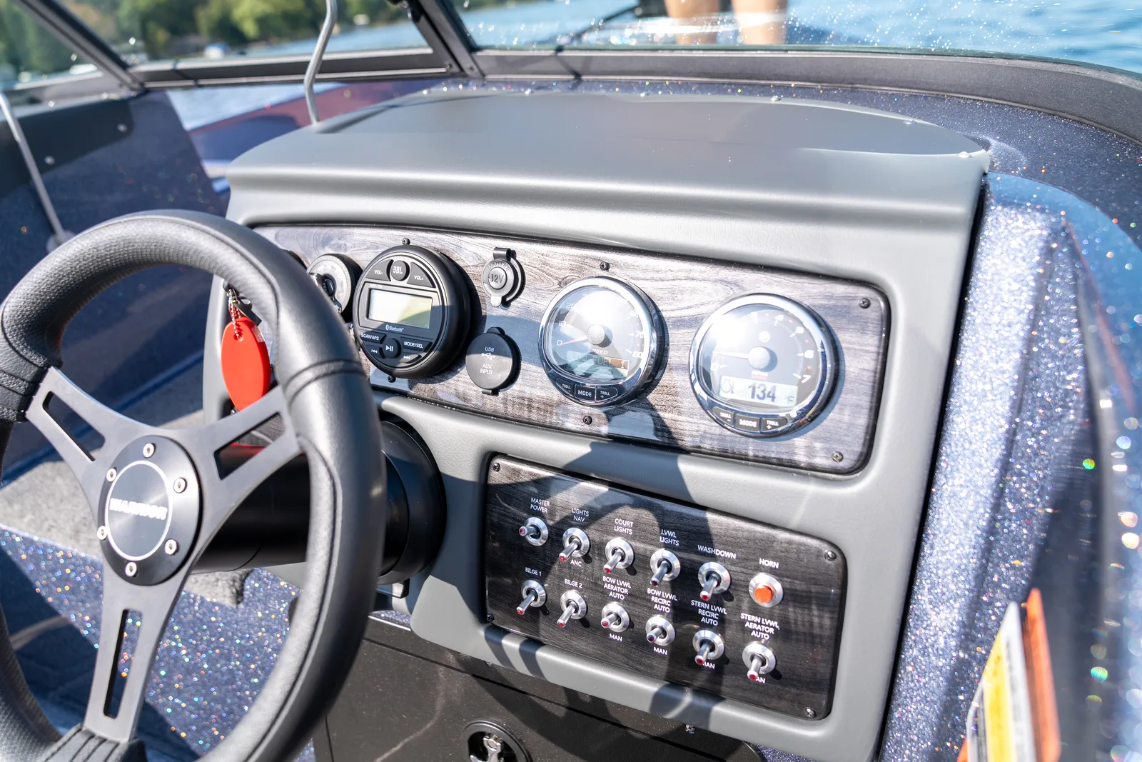 The boat helm with the steering wheel, stereo, speed and tachometer gauges and a labelled switch panel below, with the windshield and lake beyond.