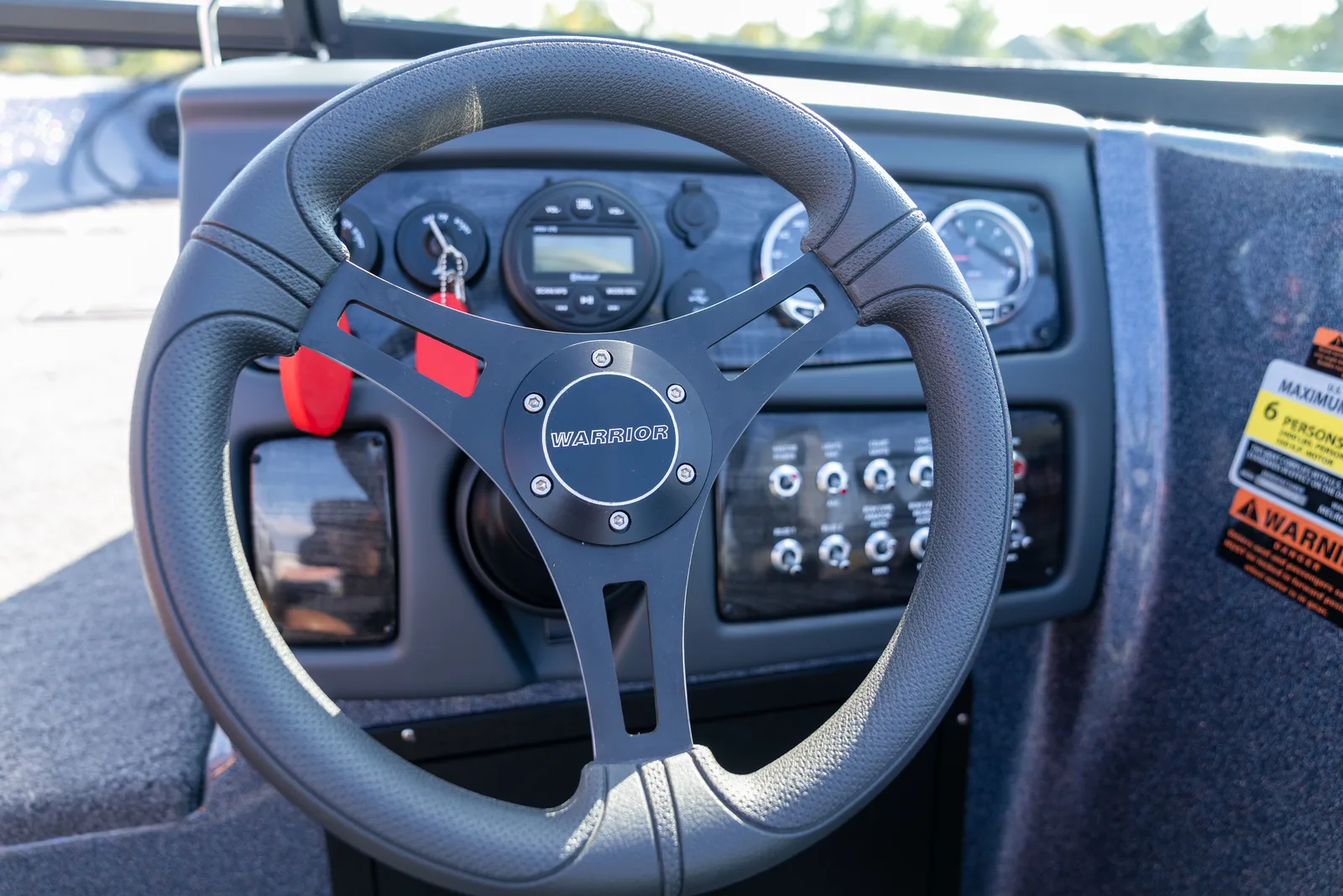 Close-up of the boat's helm showing a black steering wheel with a Warrior center cap in front of a dash with round gauges, a Bluetooth stereo, and a row of labeled rocker switches.