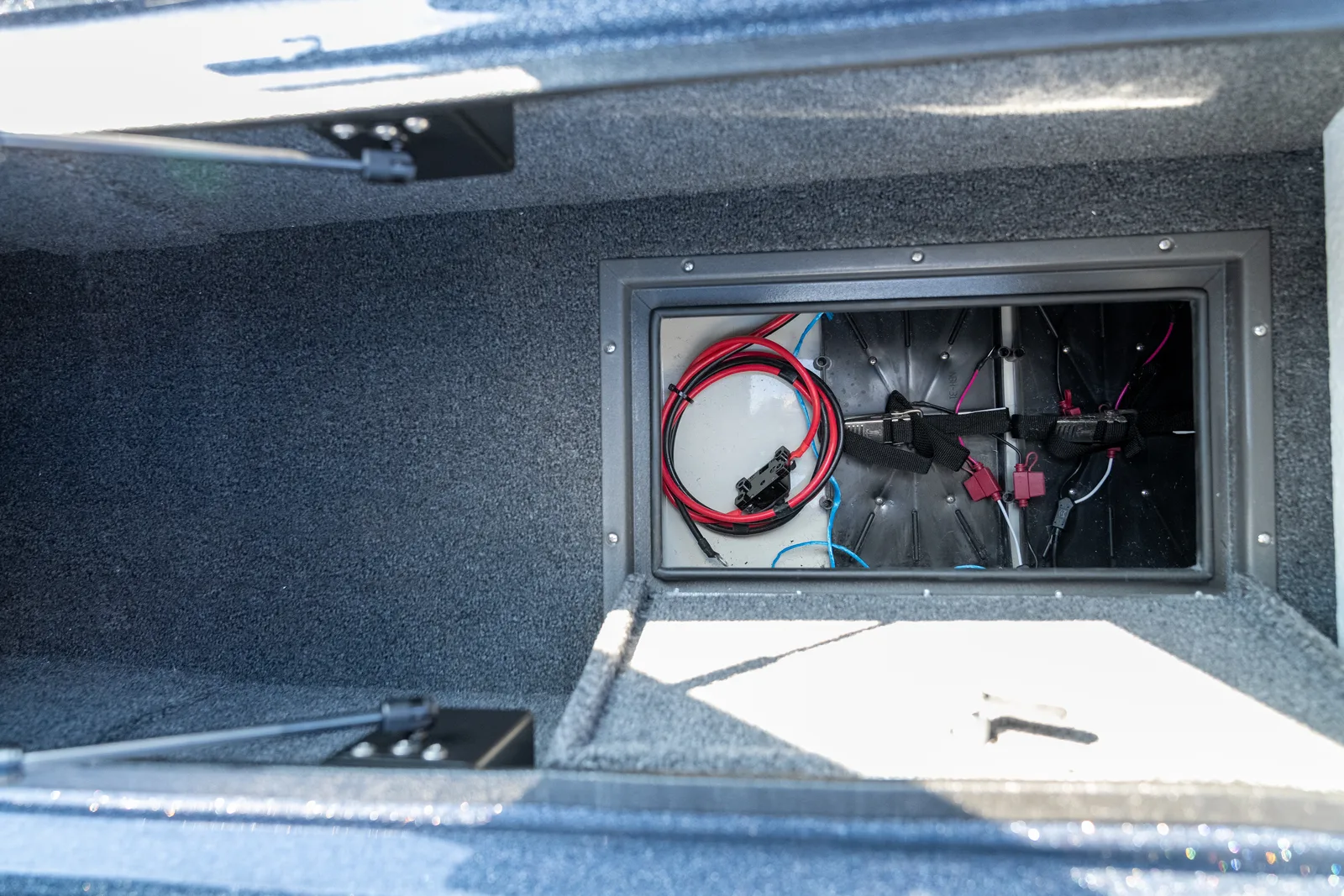 Looking down into an open carpeted storage compartment with a rectangular access panel in the floor revealing red and black battery cables, hold-down straps, and fuses.