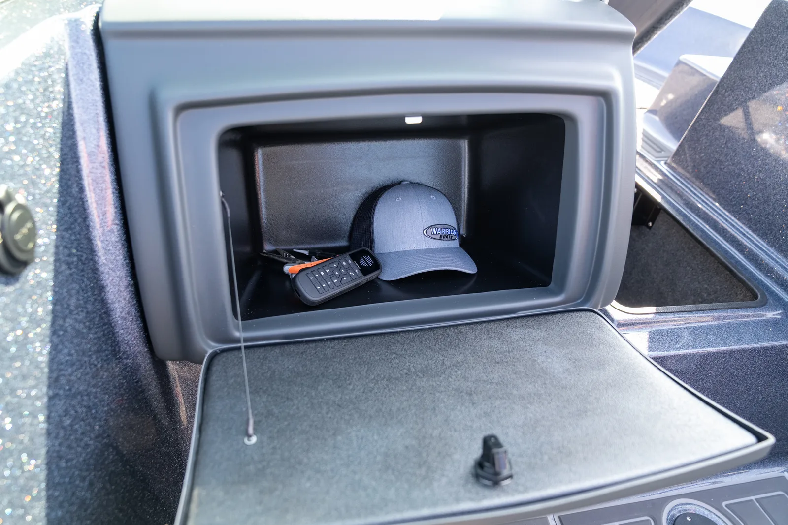An open console glove box with its door folded down, holding a grey Warrior Boats cap, a handheld remote, and an orange-handled tool.