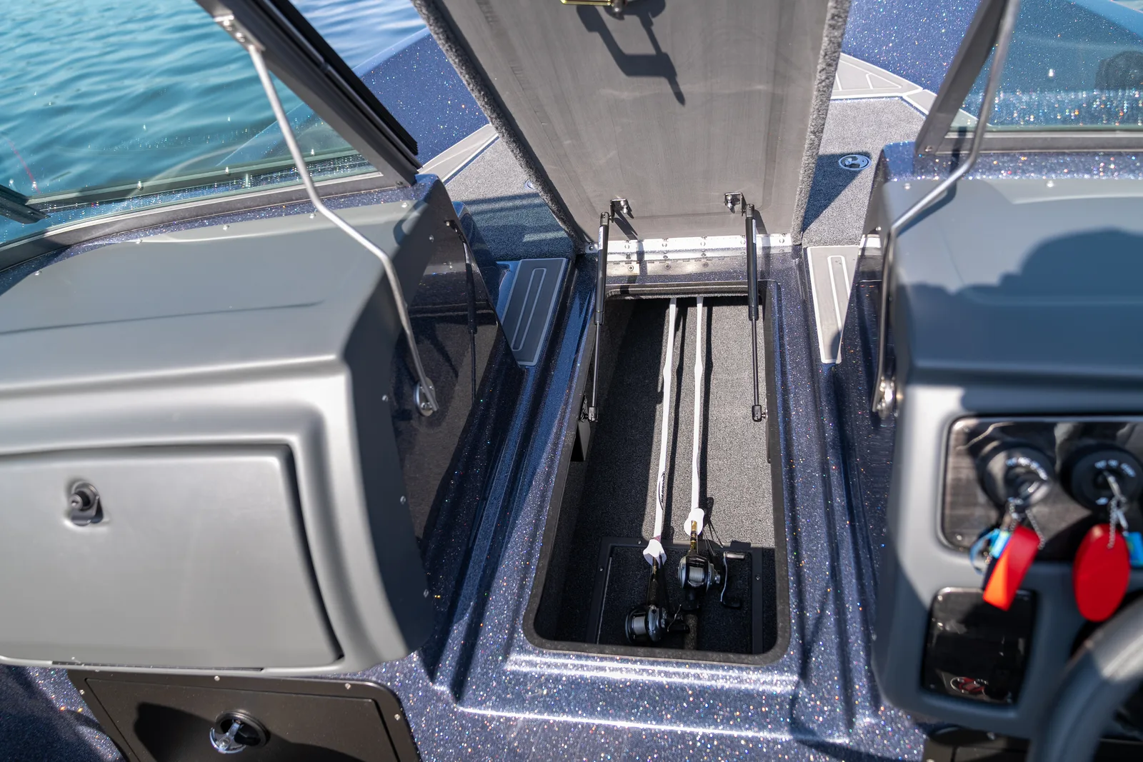 Overhead view down the cockpit walkway with the center floor rod locker open on struts, fishing rods stowed inside, flanked by the port console and the helm.