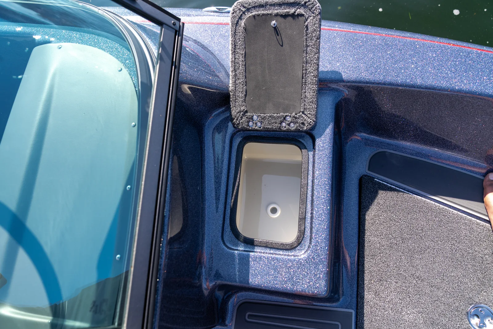 A recessed moulded pocket with a drain hole set into the boat's gunwale beside the windshield, with its hinged carpeted lid propped open above it.
