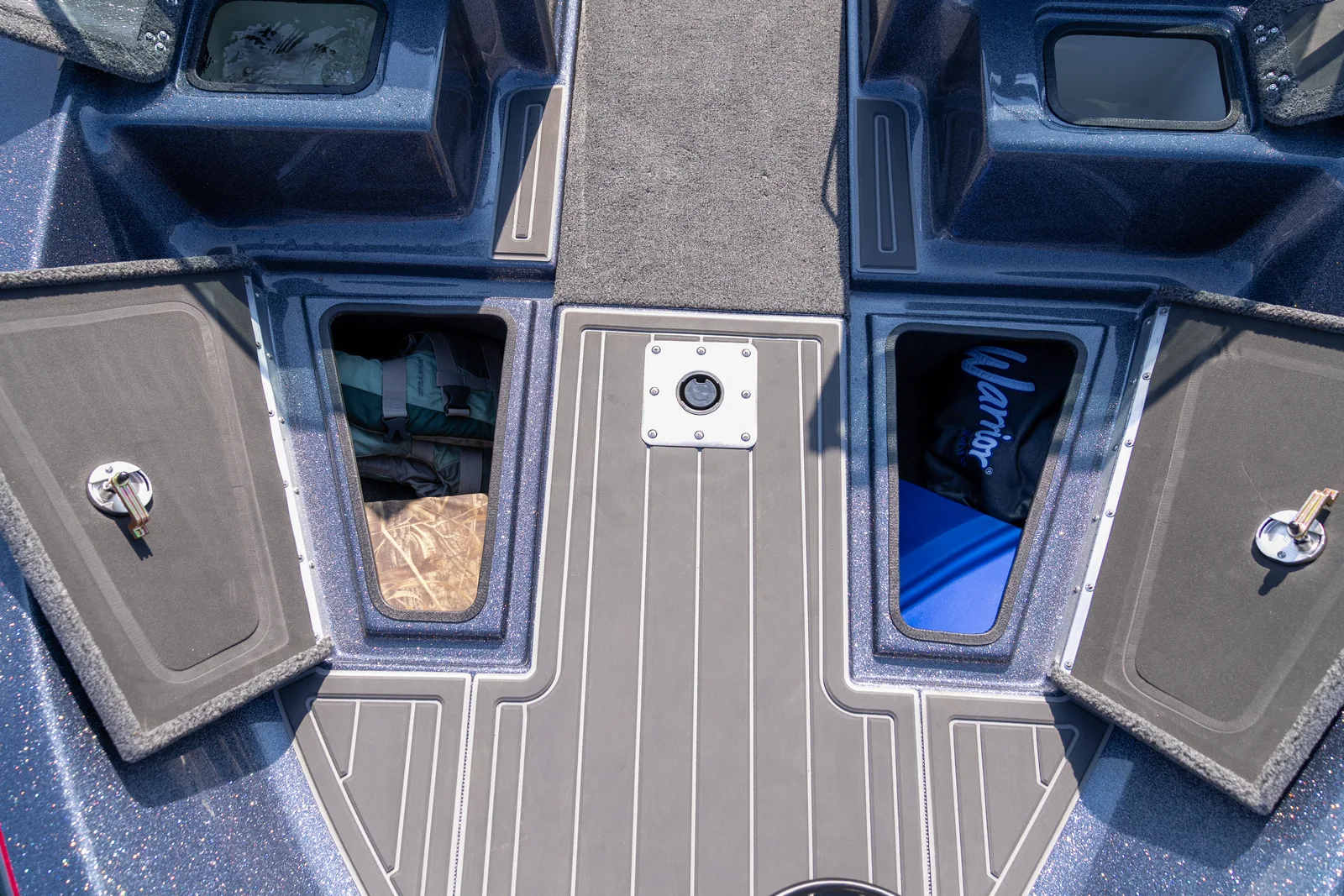 Looking down at the bow deck of a fishing boat with the carpeted storage hatches on both sides swung open to show life jackets and gear, and a flush stainless seat base set into the centre of the deck.
