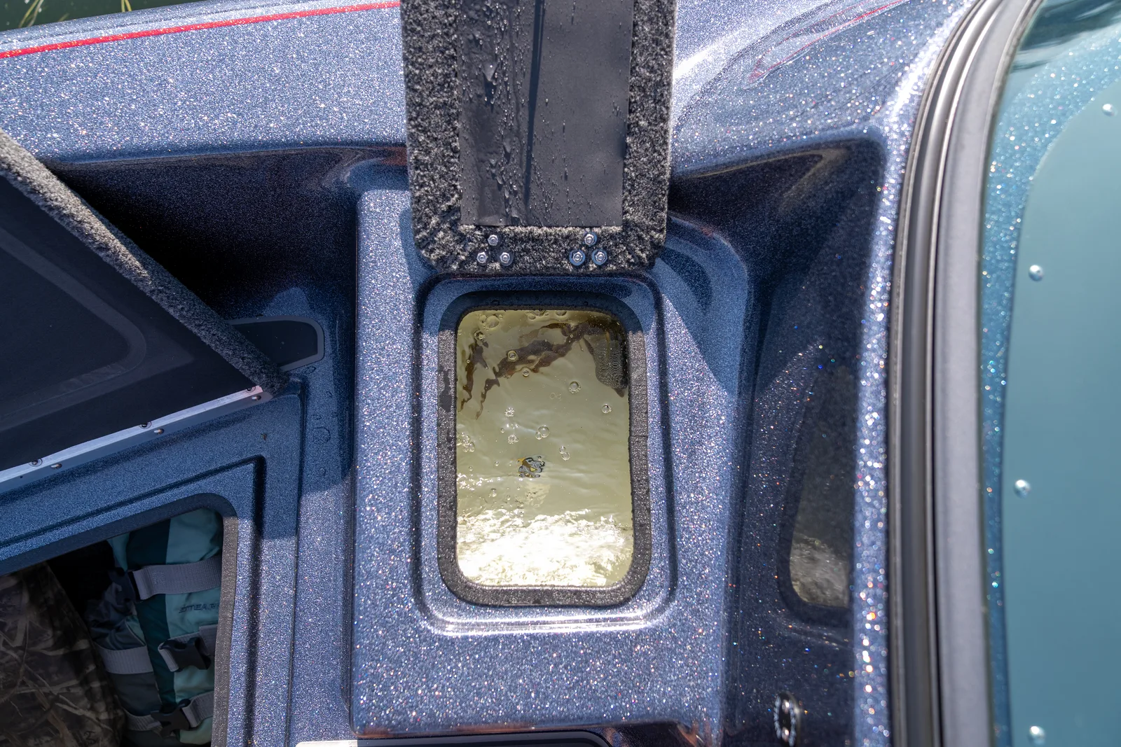An open livewell in the bow deck part-filled with aerated water, with its wet lid propped open above and a side storage compartment holding a life jacket alongside.