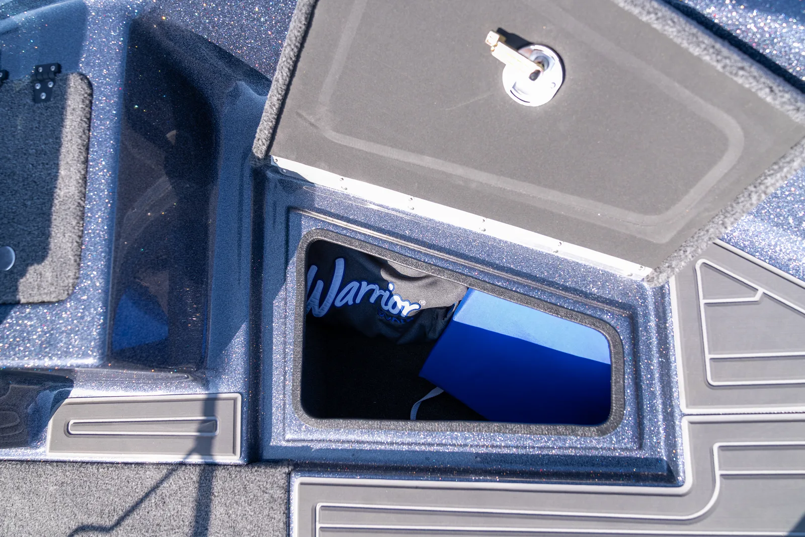 An open bow storage compartment with its carpeted lid raised on a stainless latch, holding a Warrior-branded cover bag and a blue plastic case.