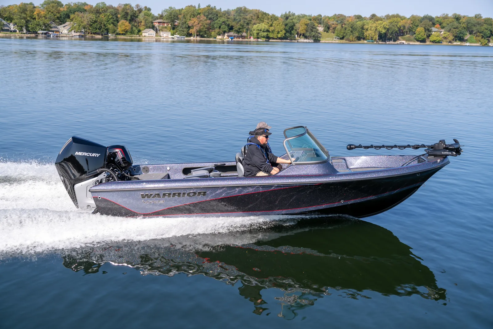 A Warrior V203 runs left to right across a blue lake with two anglers aboard, a Mercury Verado on the transom and an autumn shoreline behind.