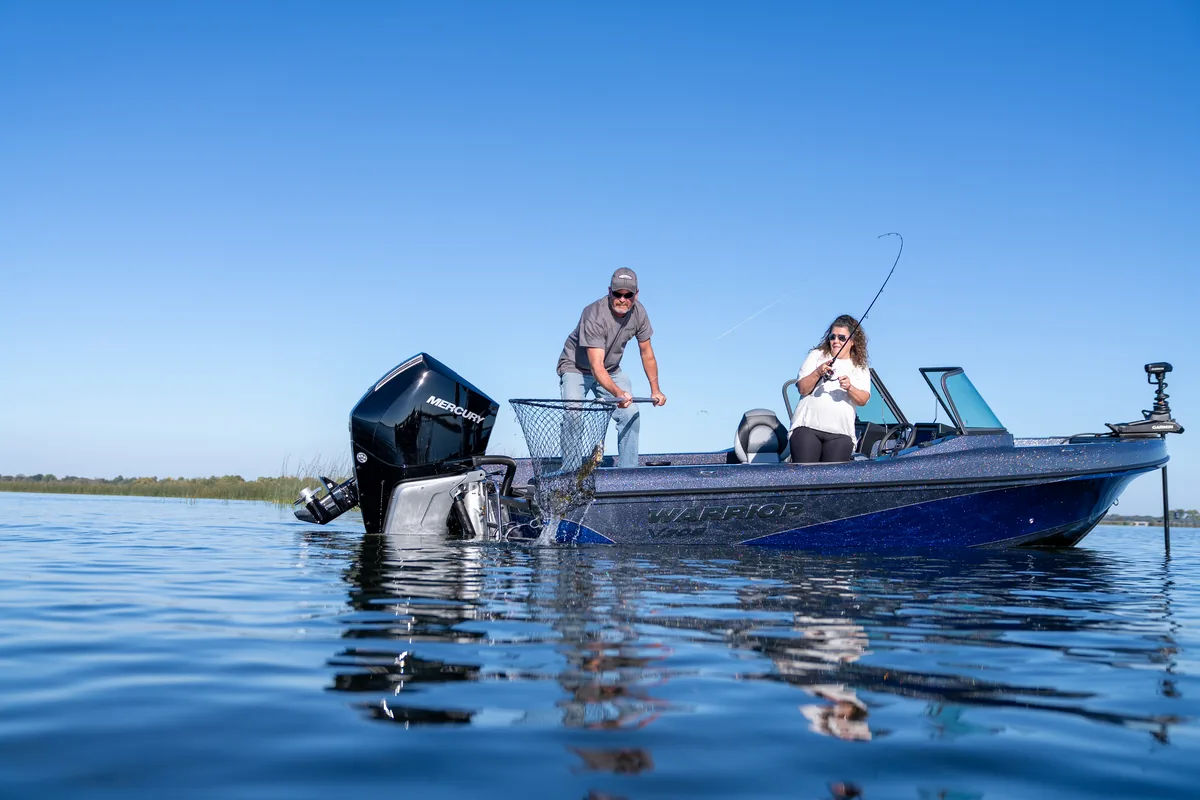 A man nets a fish over the side of a blue Warrior V208 while a woman fights another fish on rod and reel, on calm water under a clear blue sky.