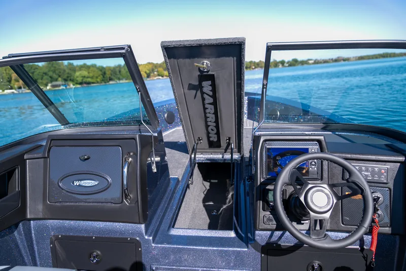 View across the helm with steering wheel, electronics display, and an open center storage hatch, with blue water beyond the windshield.