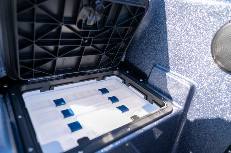 An open storage hatch in the boat side panel holding stacked clear tackle trays with blue latches.