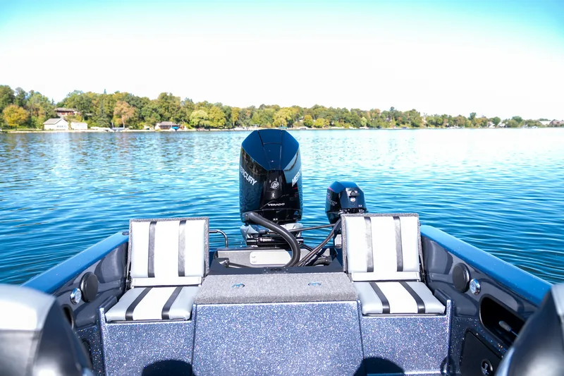 View across the rear deck of the boat toward the outboard motors with two jump seats up and a lakeshore behind.