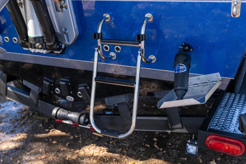 Stainless folding boarding ladder mounted on the blue transom of the boat above the trailer, with the outboard jack plate and taillight visible.