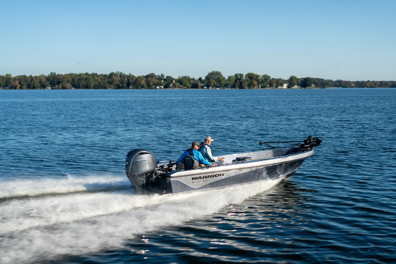 A Warrior V2090 tiller boat runs left to right across a blue lake with two anglers aboard, the Yamaha outboard at the stern and the trolling motor stowed on the bow.