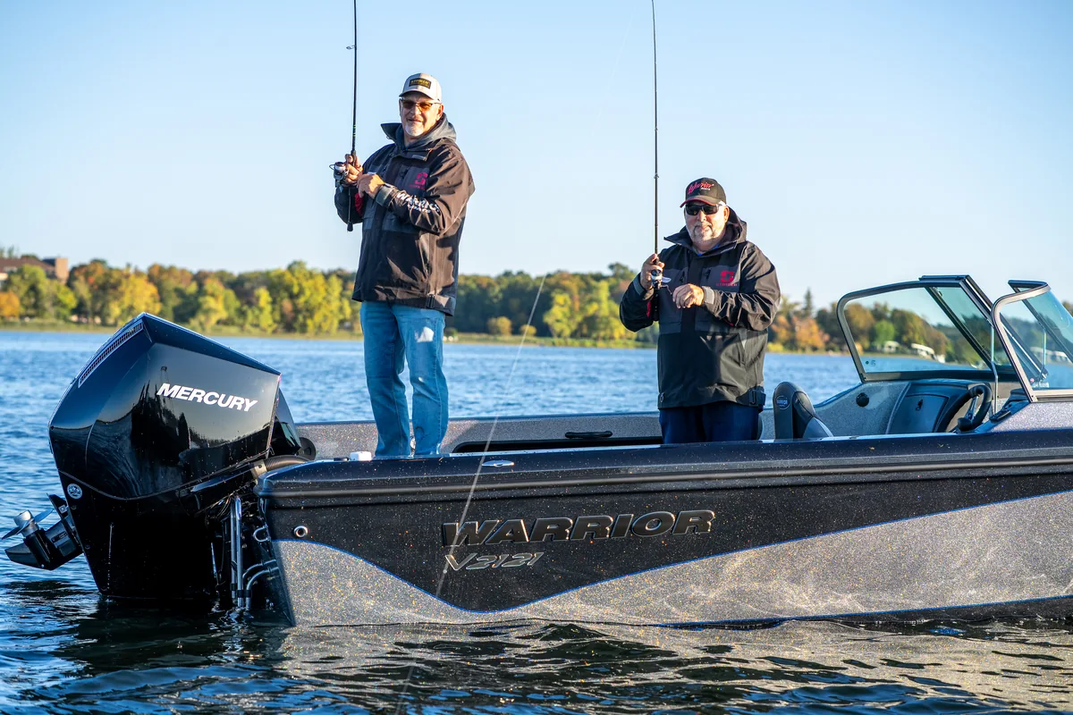 Two anglers stand casting from the deck of a Warrior V2121 on calm water with a tree-lined shore behind them.