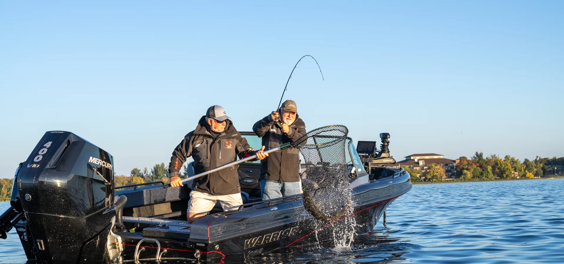 Two anglers on a black Warrior boat with a Mercury 400 outboard net a fish over the gunwale on a calm lake at golden hour.